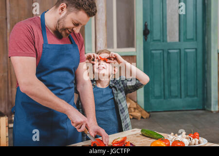 Smiling little boy looking at happy father cutting vegetables on cutting board, dad and son cooking concept Stock Photo