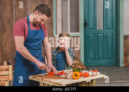 Smiling little boy looking at happy father cutting vegetables on cutting board, dad and son cooking concept Stock Photo