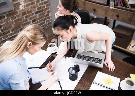 multicultural businesswomen discussing project and writing something in notepad together at workspace Stock Photo