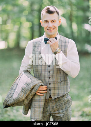 The vertical view of the handsome smiling groom touching his bowtie in the park. Stock Photo