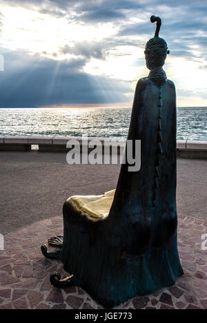 A single chair throne sculpture with head and feet looks out at the Bay of Banderas with sun bursting through clouds on the Puerto Vallarta malecon or Stock Photo