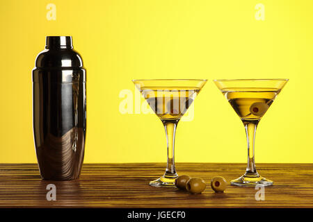 Two cocktails in martini glasses with green olives and shaker on a wooden surface against yellow background with copy space Stock Photo