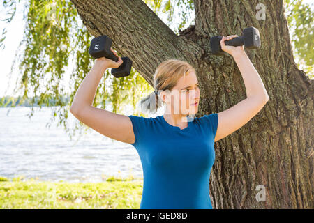 Young woman lifting dumbbells exercising 12 pound weights in green grass outdoor park by river with soft sunlight Stock Photo