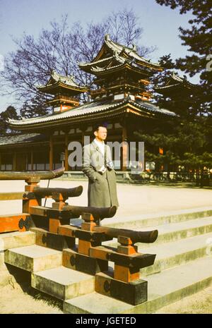 A tourist wearing a Kodak Brownie camera and an additional camera on straps around his neck walks past an ornate Japanese temple, Japan, 1955. Stock Photo