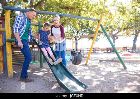 Happy parents looking at daughter playing on slide at playground Stock Photo