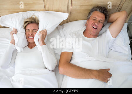 Annoyed woman covering ears with pillow while man snoring on bed in bedroom Stock Photo