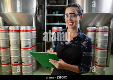 Portrait of smiling female worker holding clipboard by storage tanks at brewery Stock Photo