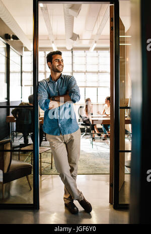Full length portrait of happy young man standing in doorway of office with his arms crossed and looking away. Male executive at office with people wor Stock Photo