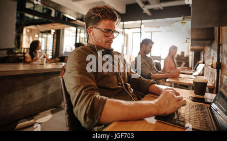 Young man sitting at cafe and using laptop computer, with people in background. Stock Photo