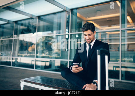 Young businessman sitting on bench with suitcase and using smart phone. Caucasian businessman waiting at airport terminal holding mobile phone. Stock Photo