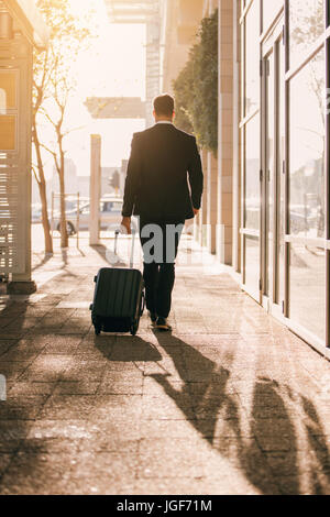 Vertical shot from behind of man walking with bag outside on city street. Young business traveler with suitcase outside airport. Stock Photo