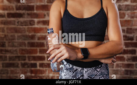 Mid section of a fit young woman holding water bottle against wall at fitness studio. Closeup of an athlete wearing a smartwatch relaxing. Stock Photo