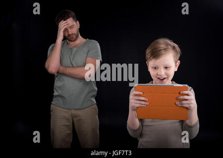 Upset father standing behind excited son using digital tablet, family problems concept Stock Photo