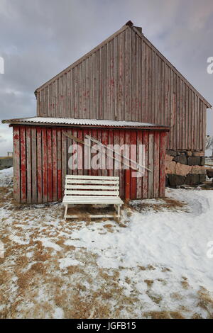 Old wooden cottage with red faded paint walls over stone slabs base-attached boatshed with white painted wooden bench on the outside. Nordmela-Andoya- Stock Photo