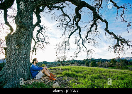 Caucasian man sitting in field leaning on tree trunk Stock Photo