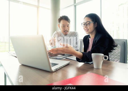 Couple of young designers working at modern office, two coworkers discussing fun project over a laptop, little team of businesspeople smiling and look Stock Photo