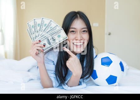 Football and money. Young Asian woman holding soccer ball and cash in bedroom at home. Competition lottery gambling concept idea Stock Photo