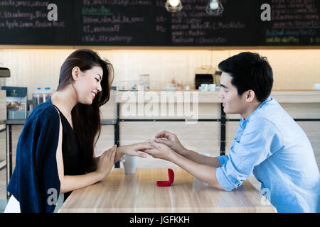 Asian handsome man putting wedding ring on and proposing to his beautiful woman in cafe. Love, anniversary, surprise, people and holidays concept