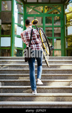 Casual businessman going to work by bicycle. He is carrying the bike upstairs to the entrance of the office building. Rear view. Stock Photo