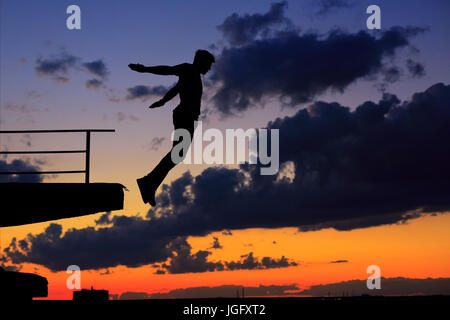 Silhouette of a man against a background of clouds and golden sunset. He jumps from the roof edge. Stock Photo