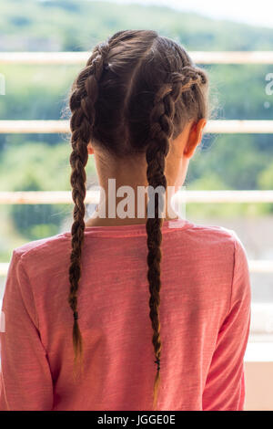 Girl with Dutch plaits looking out of window from behind. Young child with brown hair braided in a pink top Stock Photo