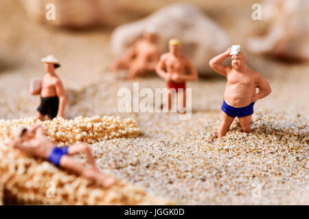some different miniature men wearing swimsuit relaxing next to some seashells and a starfish on the sand of the beach Stock Photo