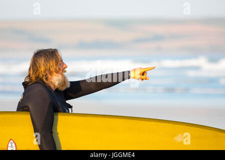 A mature male surfer with long beard holding a surf board and pointing towards the sea at the popular surfing beach of Polzeath, Cornwall Stock Photo