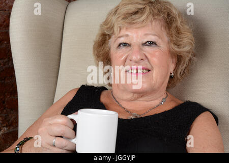 Portrait of an old woman drinking coffee and sitting on couch. Indoors. Stock Photo
