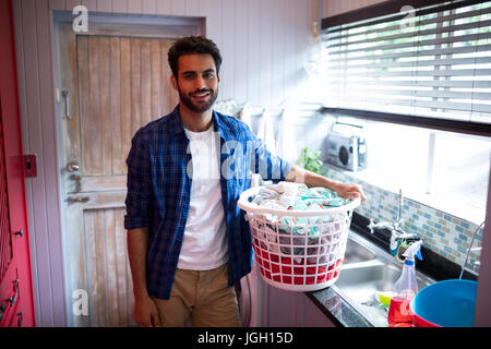 Portrait of young man holding laundry basket while standing by sink at home Stock Photo