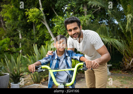 Portrait of father with son sitting on bicycle in yard Stock Photo