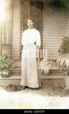 Full-body portrait of African American woman standing in front of a porch, wearing a white dress, with her hat on the ground, potted plants by her side, with a serious facial expression, 1920. Note: Image has been digitally colorized using a modern process. Colors may not be period-accurate. Stock Photo