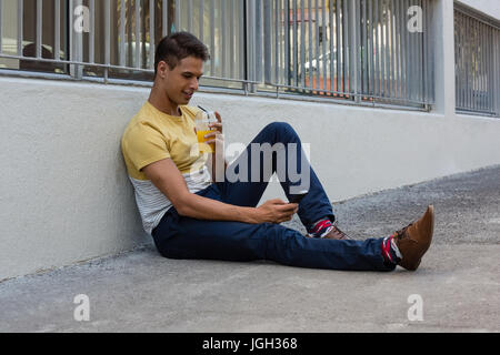 Young man having drink while using mobile phone on sidewalk by wall Stock Photo