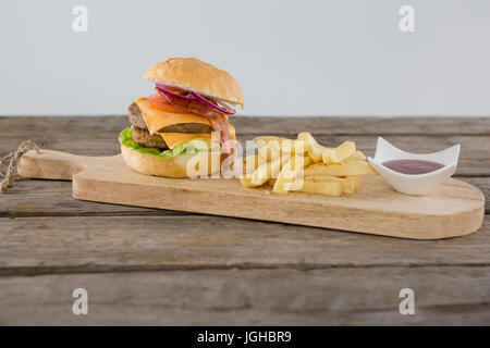 Burger with French fries and sauce on cutting board against wall Stock Photo