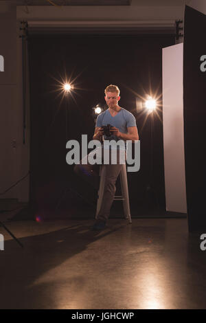 Full length of man adjusting camera while sitting on stool against backdrop at studio Stock Photo
