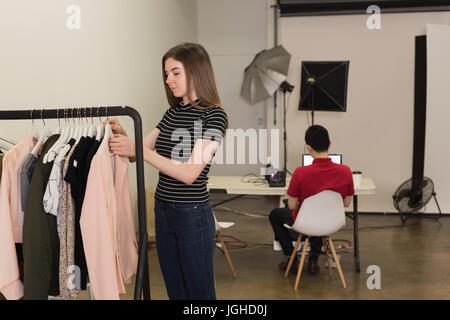 Young model choosing clothes on rack with photographer sitting on chair at studio Stock Photo