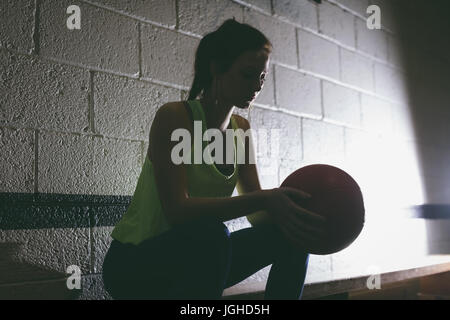 Female player holding basketball by wall while sitting on bench Stock Photo