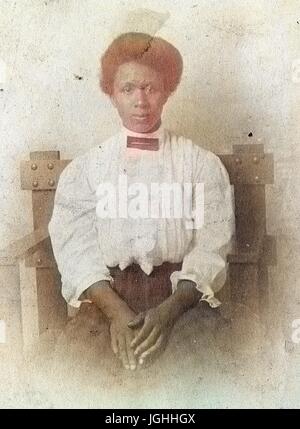 Waist up portrait of seated African American woman, wearing a white blouse and checkered dress, with a serious facial expression, hands rested on lap, 1920. Note: Image has been digitally colorized using a modern process. Colors may not be period-accurate. Stock Photo