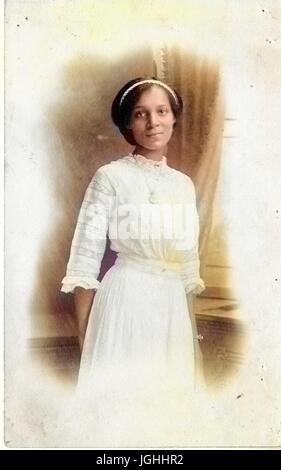 Young African-American woman in white dress, hands behind her back, with a serious expression, wearing a white headband, 1915. Note: Image has been digitally colorized using a modern process. Colors may not be period-accurate. Stock Photo