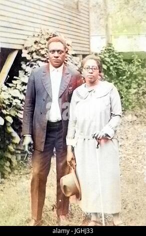 African American husband and wife standing next to each other in front of a wood building and shrubbery, the woman is wearing a light-colored shirt and long skirt and is holding a hat in one hand and a cane in the other, the man is wearing a full dark-colored suit with a light colored shirt underneath and he is holding a cigar in one hand, 1950. Note: Image has been digitally colorized using a modern process. Colors may not be period-accurate. Stock Photo