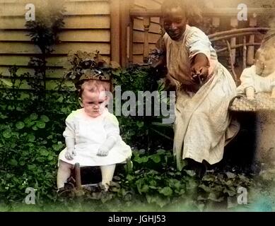 African-American nanny in a garden beside a home, wearing a plain dress and holding out a flower, with two young Caucasian babies, 1910. Note: Image has been digitally colorized using a modern process. Colors may not be period-accurate. Stock Photo