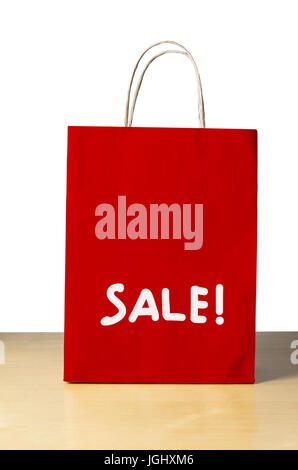 A red carrier bag on a light wood table agianst a white background with the word 'SALE!' written on the lower half in white. Stock Photo