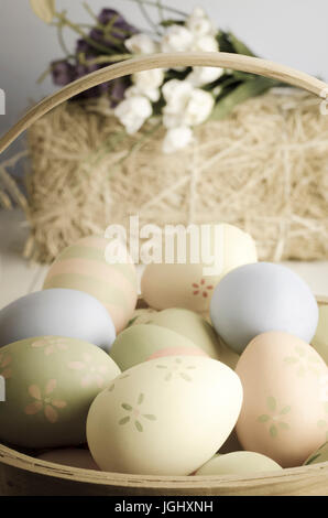 A selection of decorated Easter eggs in a wicker basket on a table, with straw bale and crocuses in the background. Stock Photo