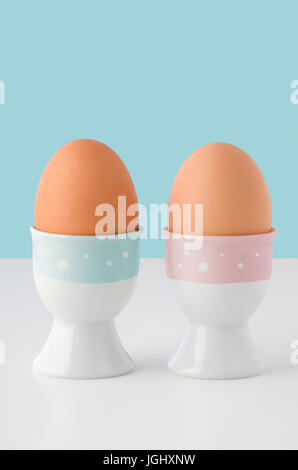 Two egg cups, decorated with pink and blue stripes with white spots and holding brown eggs on white table with light blue background.  Copy space abov Stock Photo