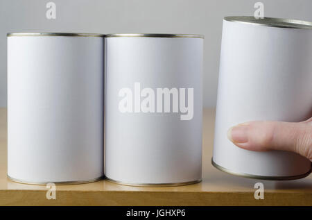 Conceptual image of three tin cans with blank white paper labels on a shelf, with middle aged female hand reaching in from right of frame, picking one Stock Photo