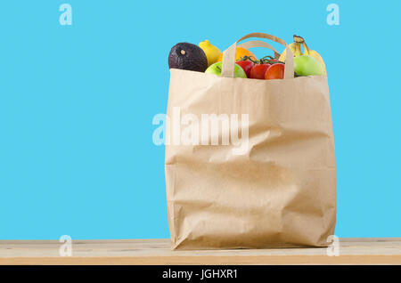 A brown paper shopping bag, filled to the top with varieties of fruit, on a light wood surface.  Isolated on a turquoise blue background. Stock Photo
