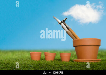 Plant pot containing potting tools, next to a row of three tiny seedling pots on grass with bright blue sky and white fluffy cloud in bacikground. Stock Photo
