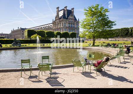 The Tuileries garden in Paris, France, by a sunny morning with a man resting on a metal lawn chair near a water basin and the Louvre palace. Stock Photo