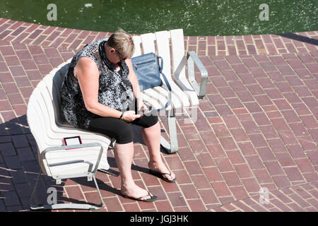 A flip-flop wearing middle-aged woman sitting on a bench at the Barbican Centre in London whilst absorbed in texting on her phone Stock Photo