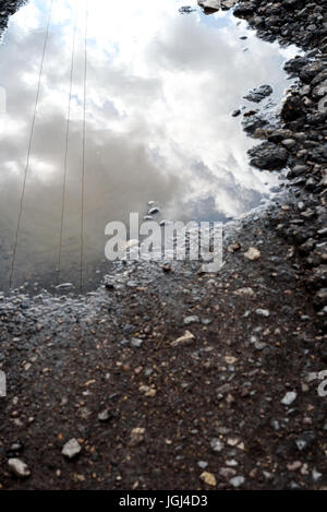 Cracked and broken asphalt pothole on the surface of a crumbling road. Rain water puddle collects in the cracked hole reflecting the sky. Stock Photo