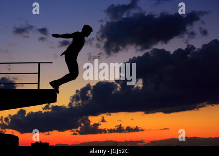 Silhouette of a man against a background of clouds and golden sunset. He jumps from the edge of the roof. Stock Photo